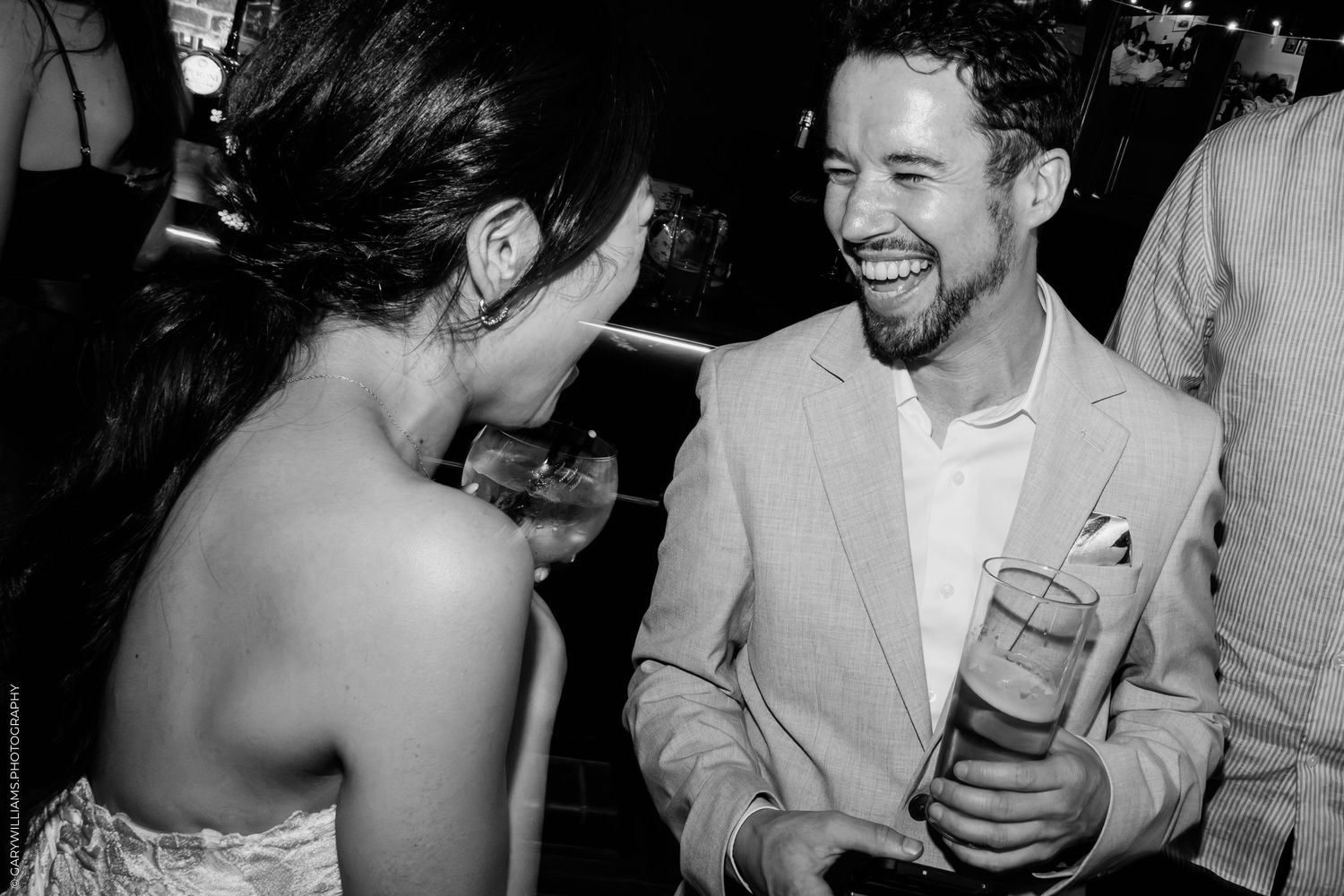 Black and white photo of a laughing man in a suit holding a drink at a lively party event.
