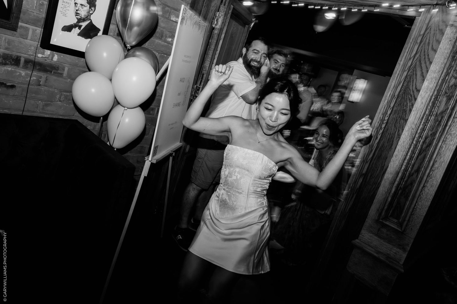 Black and white photo of a bride in a short white dress dancing joyfully at her wedding reception with guests celebrating.