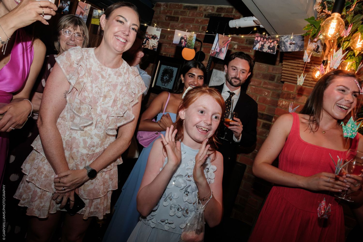 Women and a young girl dancing and celebrating at a festive indoor party with string lights and brick walls.