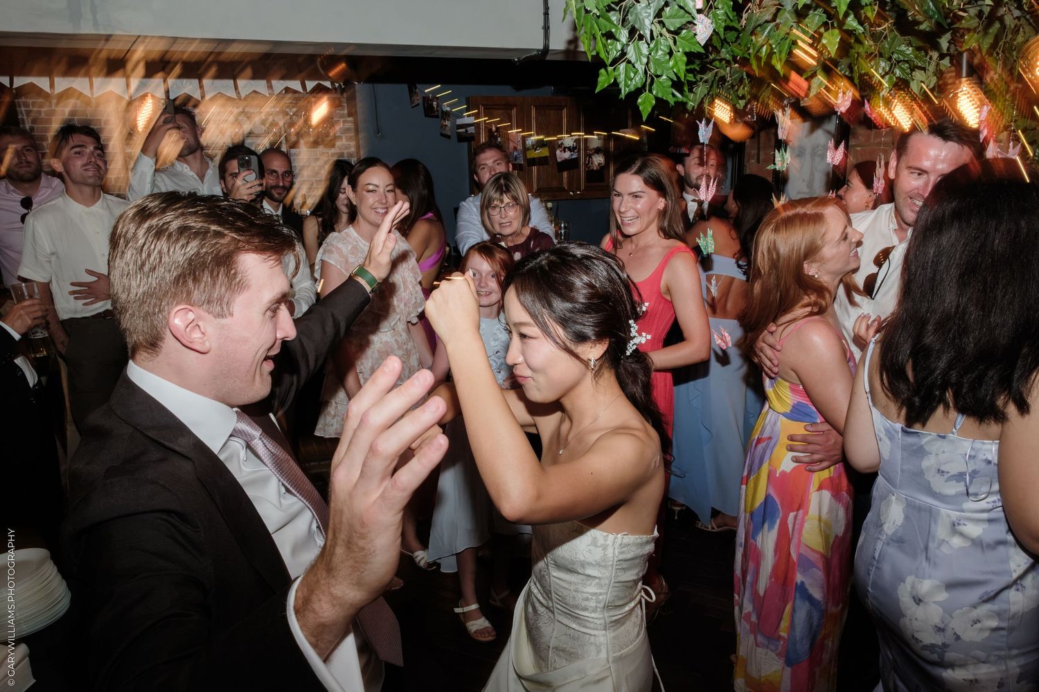 Bride and groom dancing joyfully with wedding guests at a lively indoor reception party.