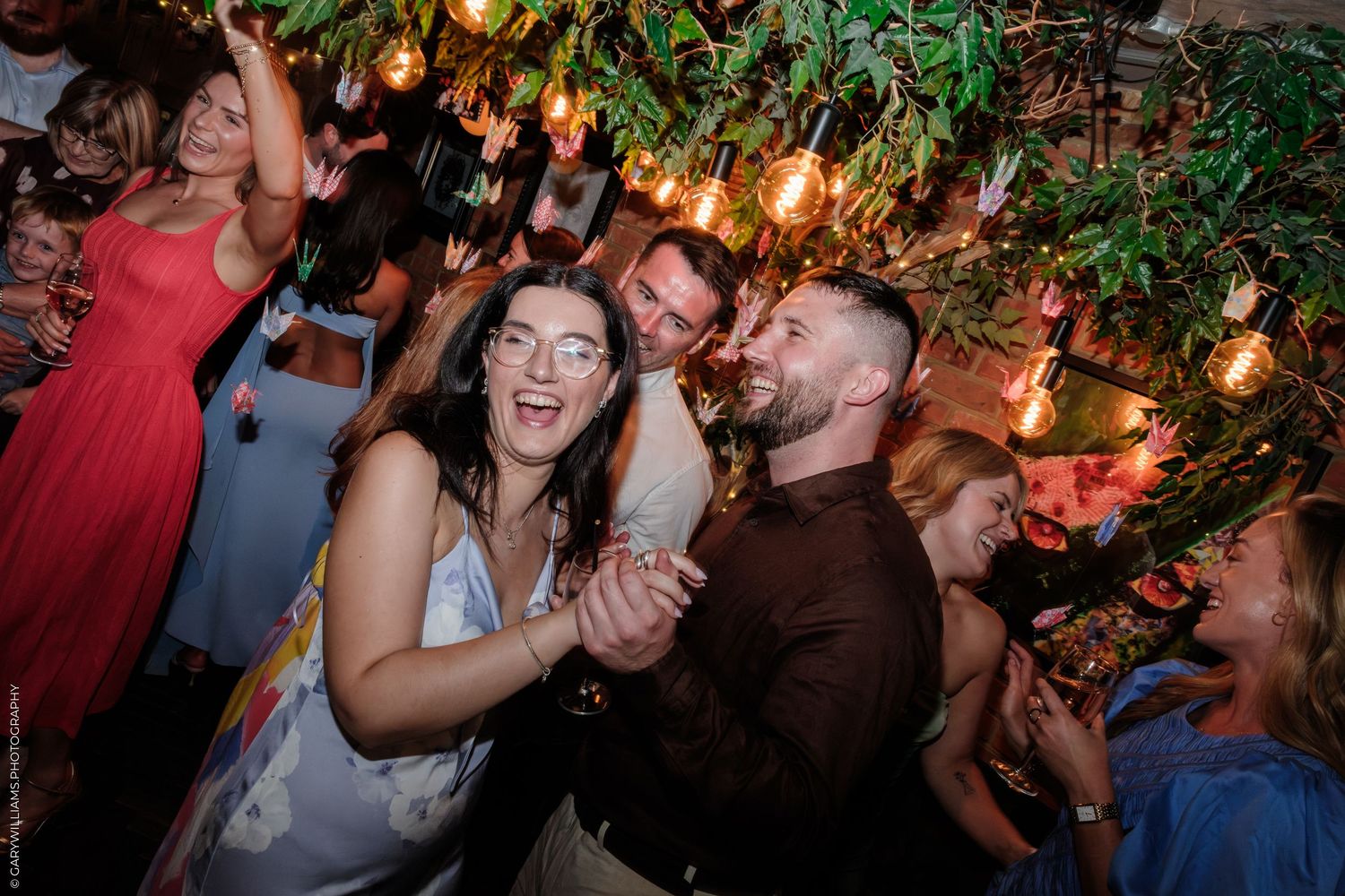 Couple dancing joyfully at a festive indoor party with warm Edison bulb lights and lush green foliage overhead.