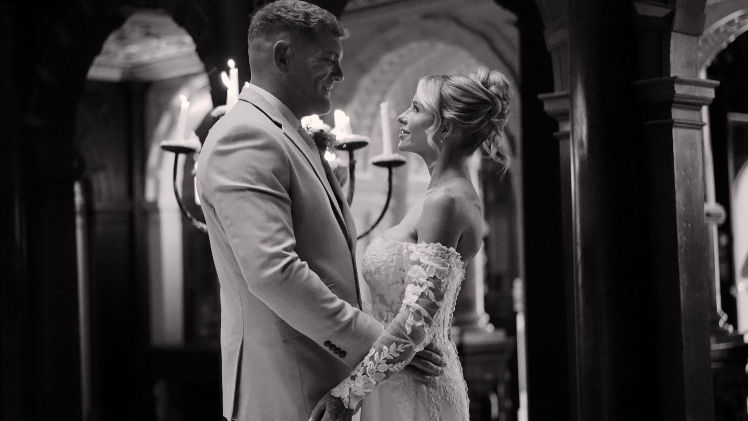 Groom giving a speech at wedding reception in elegant hall with ornate fireplace and bride seated at decorated table.