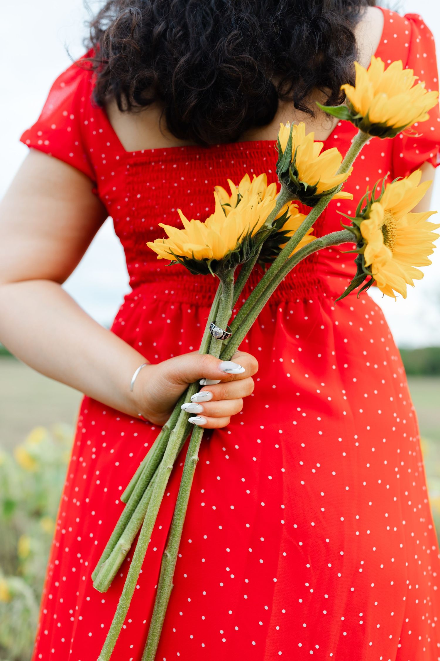 Woman in red polka dot dress holding yellow sunflowers behind her back in a field.