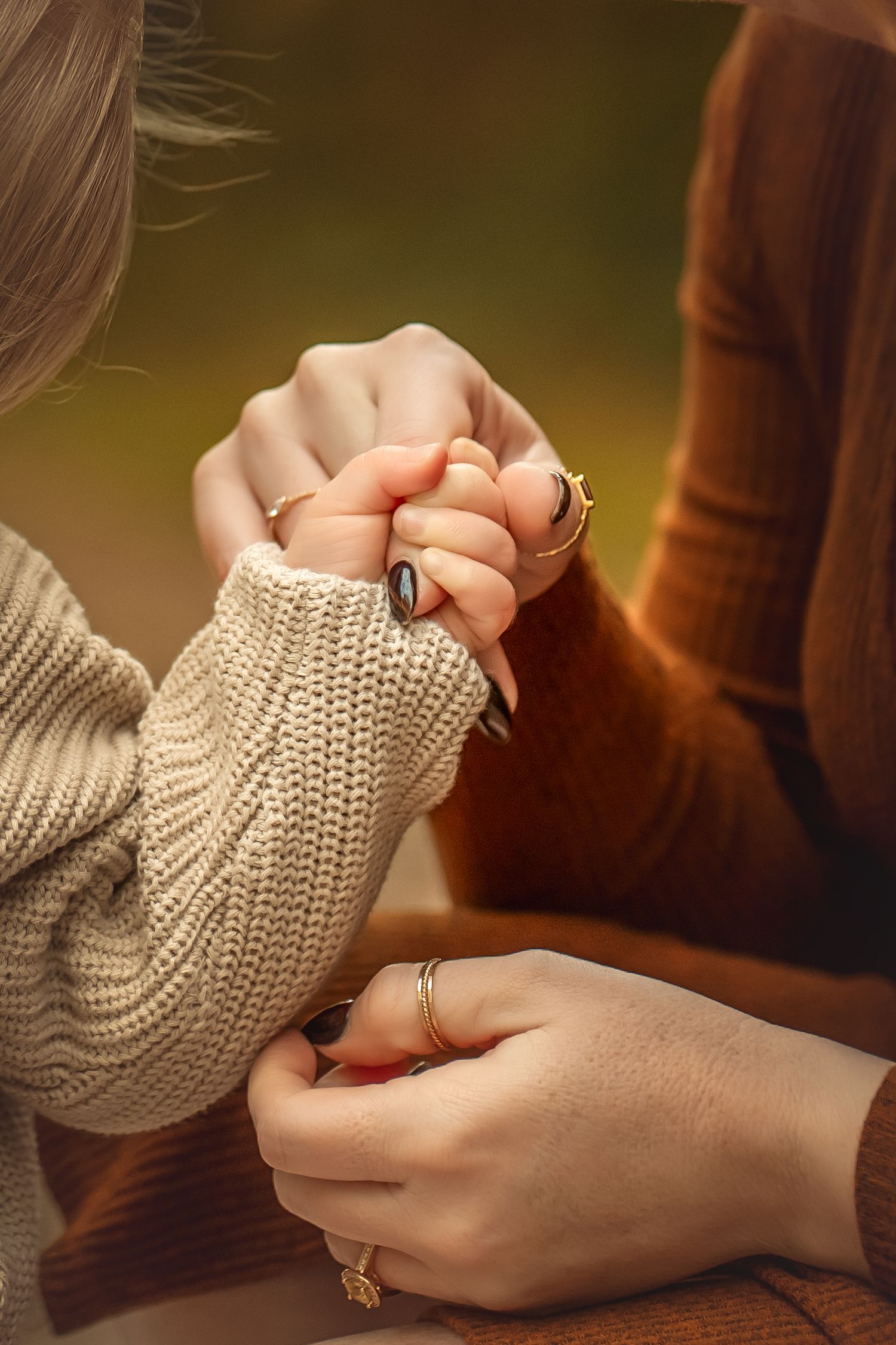Two women holding hands warmly, wearing knit sweaters and gold rings, in a cozy autumn outdoor setting.