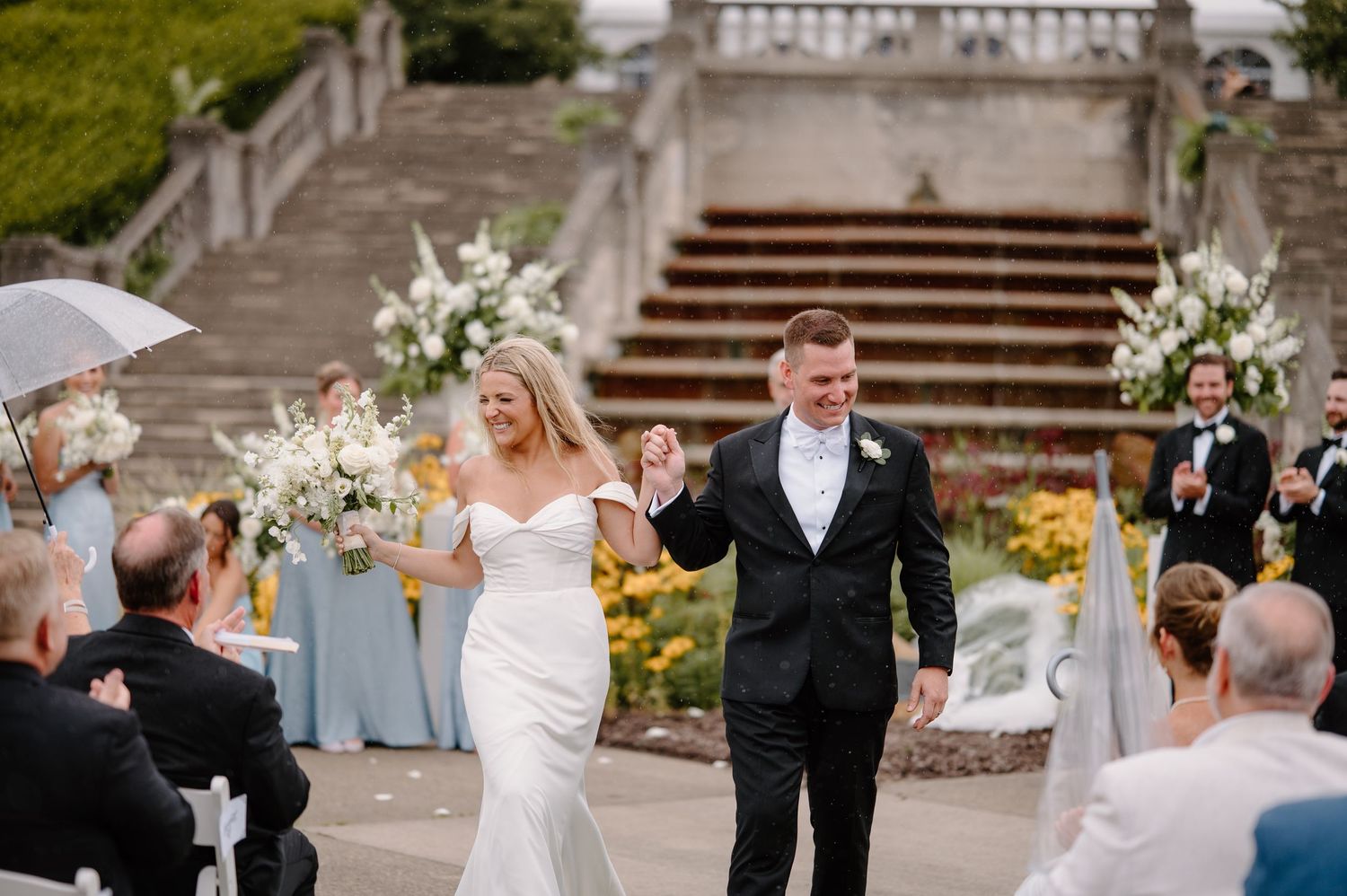 Bride in feathered dress and groom in navy suit smile holding champagne at elegant wedding reception.