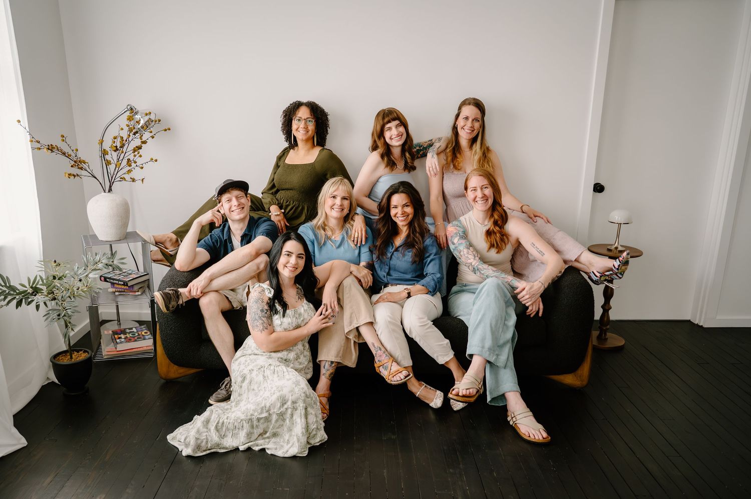 Group of nine women posing together on a dark couch, smiling warmly in a bright studio setting.