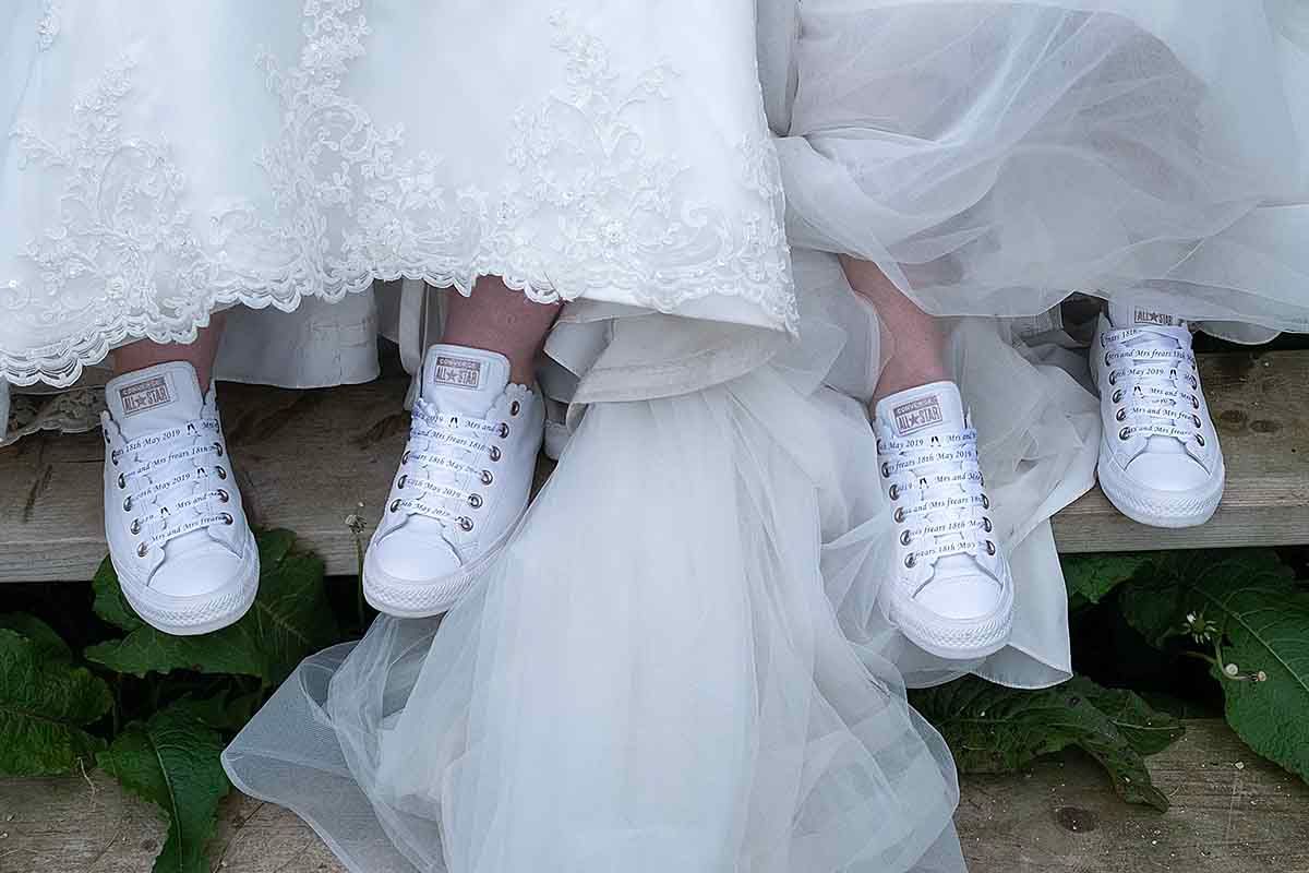 Two brides in white wedding gowns wearing matching white Converse sneakers, sitting side by side on wooden steps.