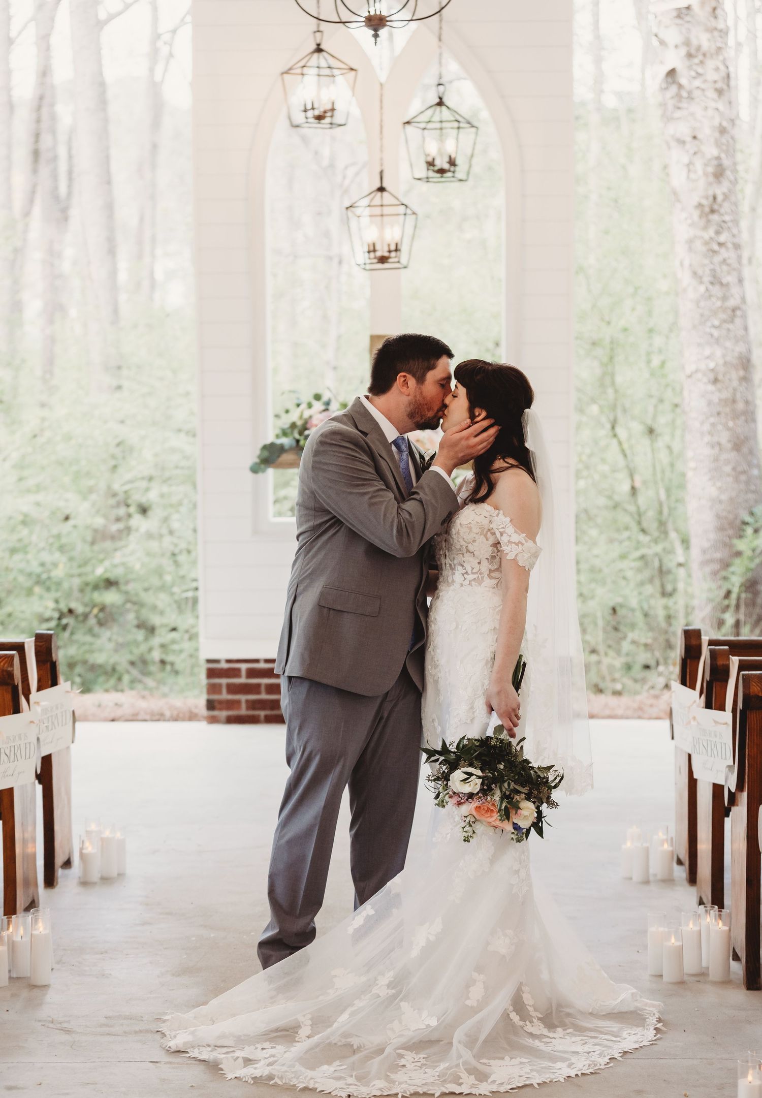 Bride and groom share first kiss in elegant chapel aisle, bride in lace gown holding floral bouquet, groom in gray suit.