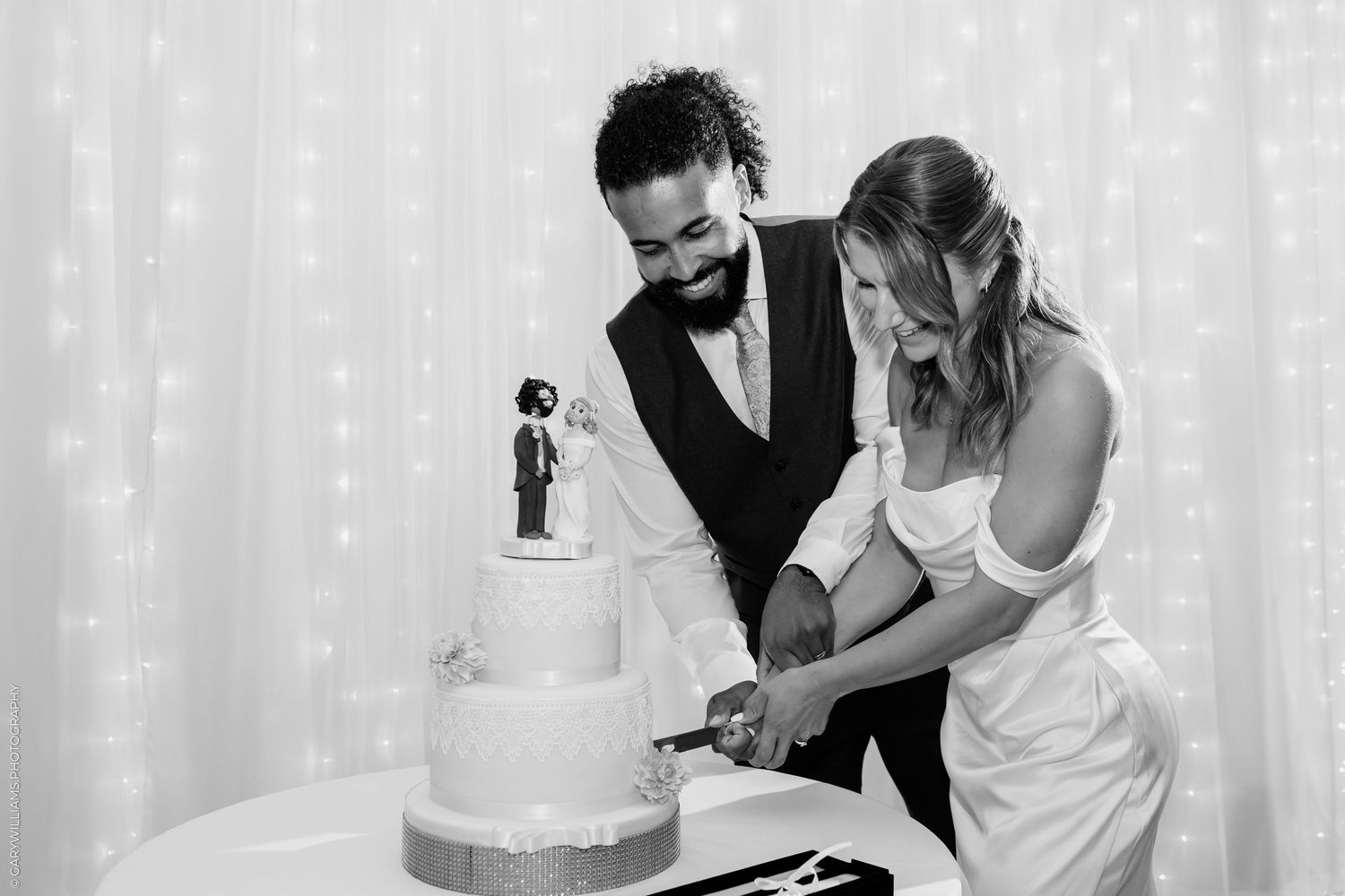Black and white photo of bride and groom cutting their tiered wedding cake together at their reception.