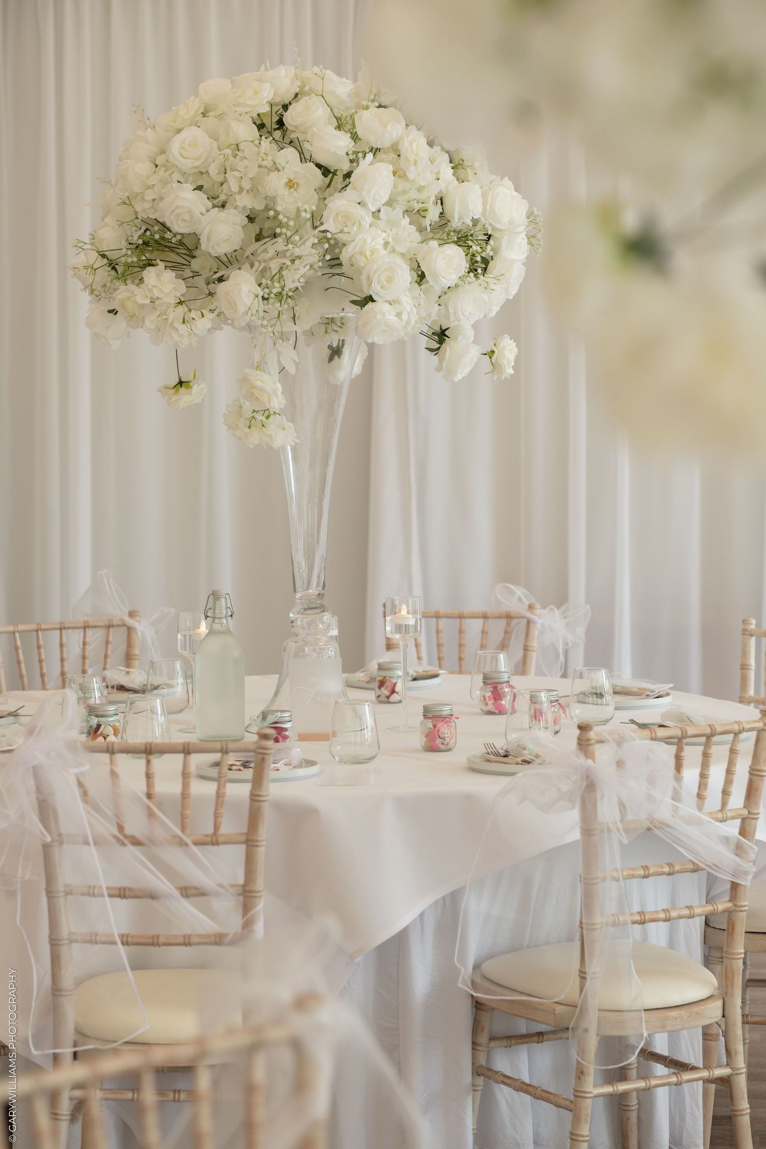 Elegant wedding reception table with tall white floral centerpiece, chiavari chairs, and white linen in a draped venue.