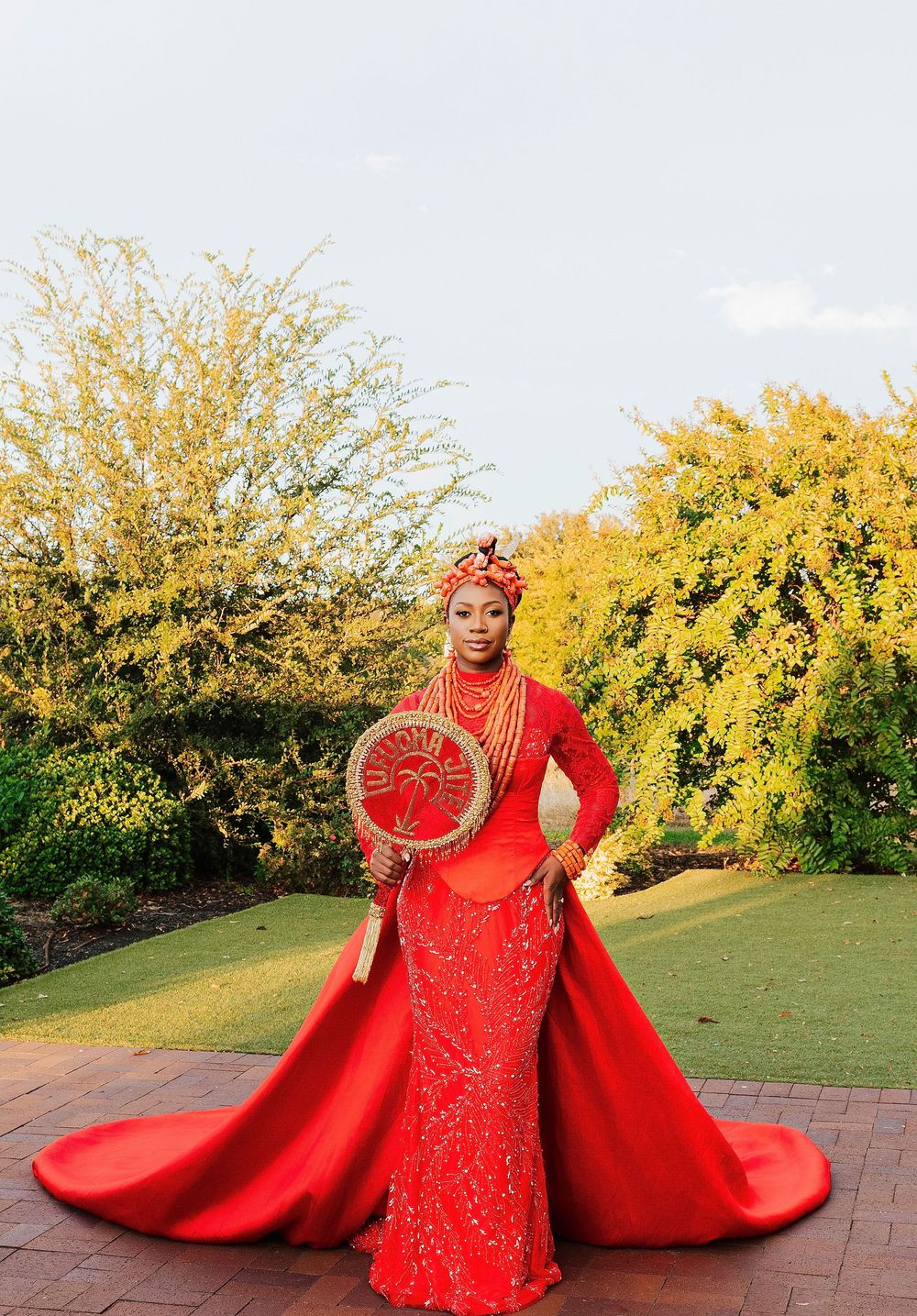 Woman in red Nigerian traditional bridal attire with gold jewelry, headwrap, and decorative fan, posing outdoors.