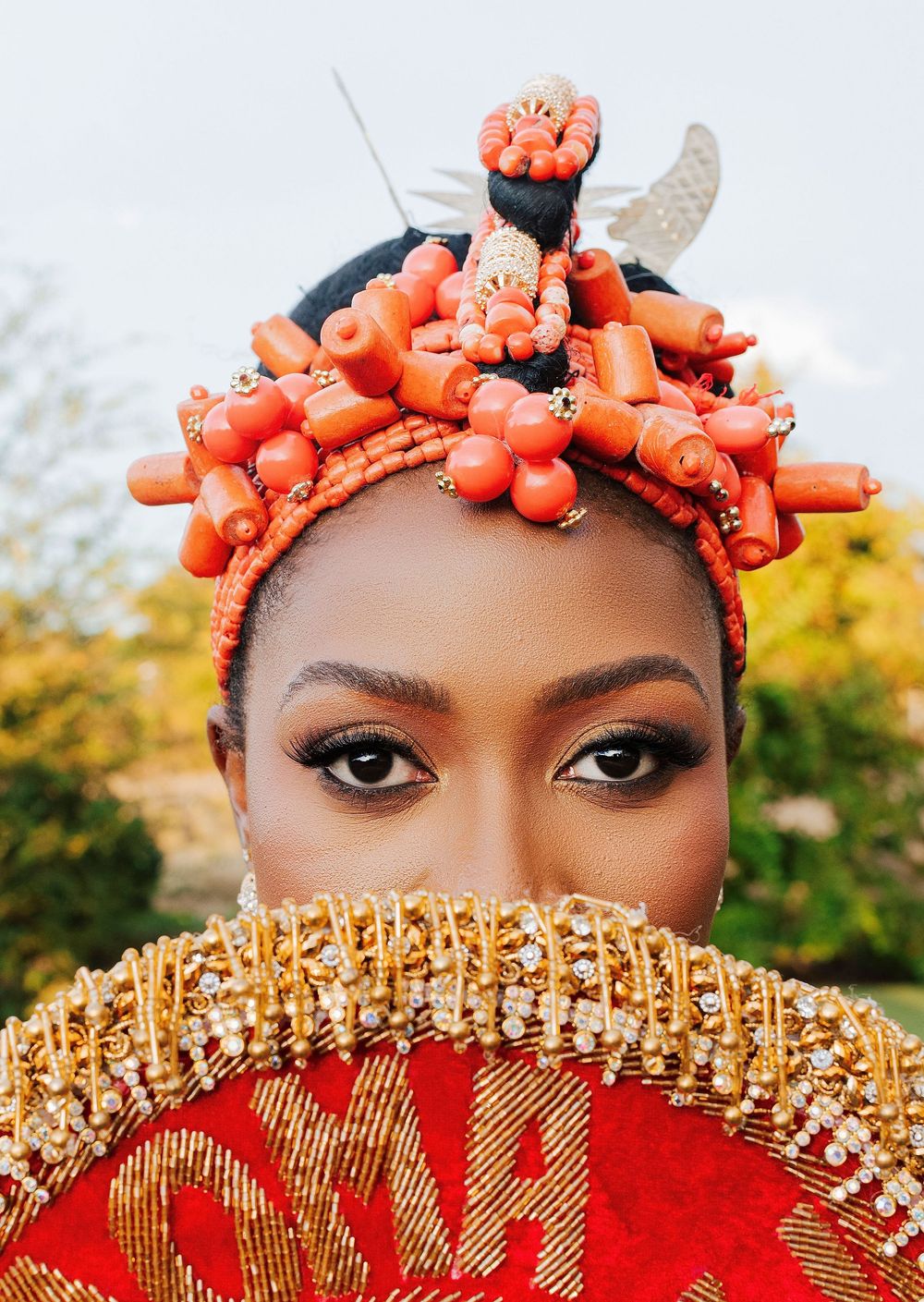 Woman in traditional Nigerian coral beaded headdress and gold fan, showcasing vibrant cultural bridal attire.