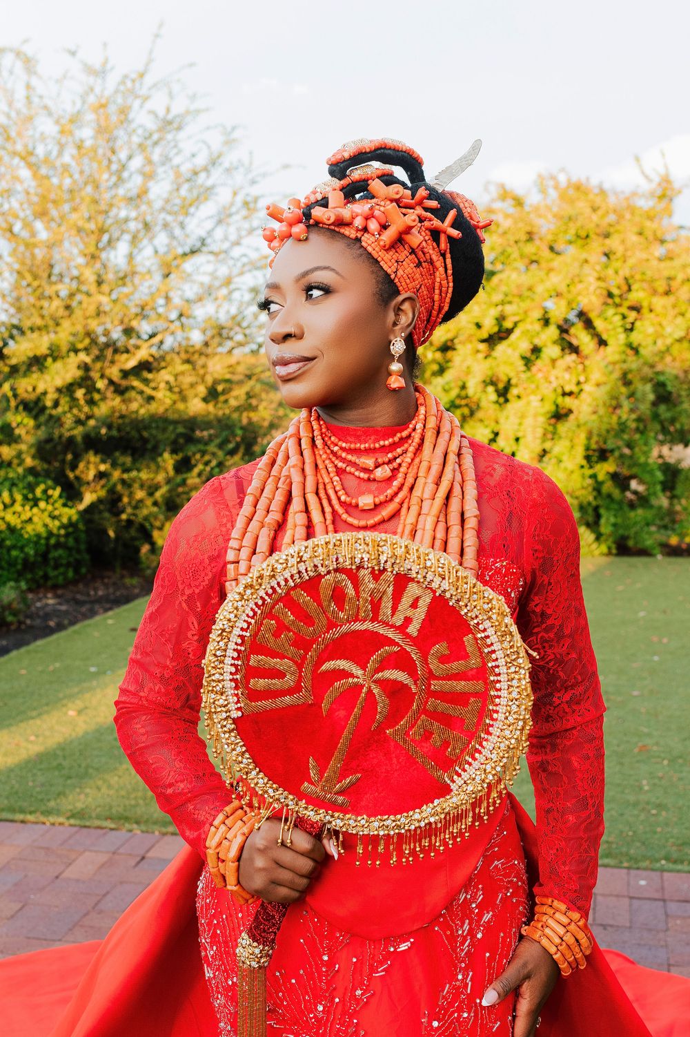 Woman in traditional Nigerian Urhobo bridal attire with red coral beads, lace dress, and decorative fan outdoors.