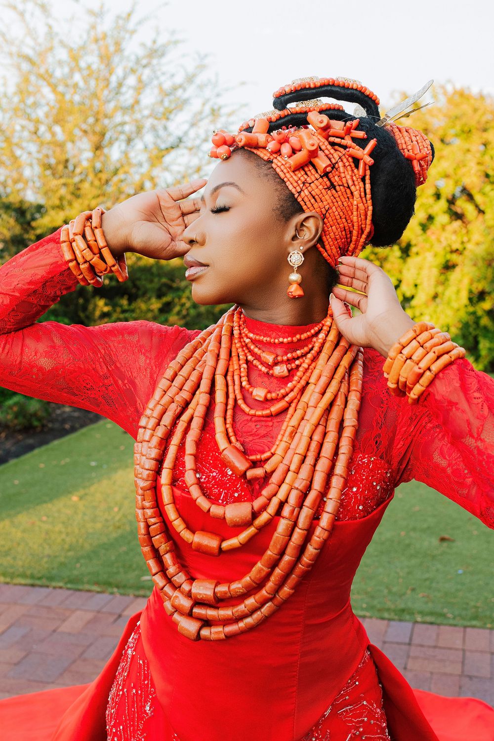 Woman in traditional Nigerian coral beaded jewelry and red attire poses gracefully outdoors.