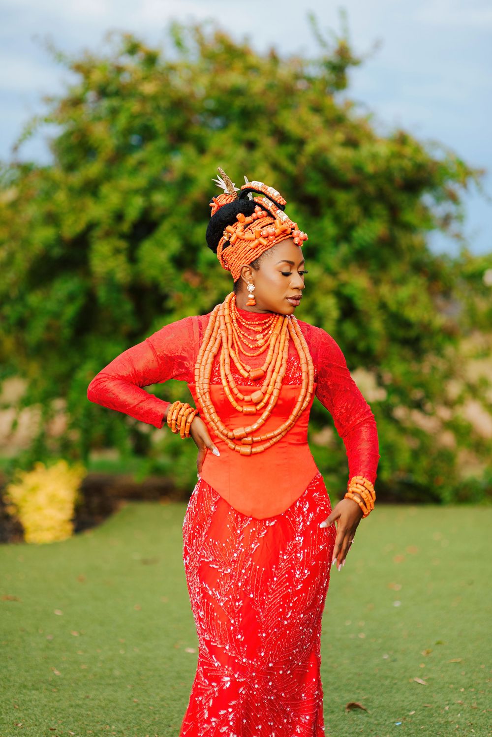 Woman in traditional Nigerian attire with orange coral beads, red lace dress, and ornate headpiece poses outdoors.