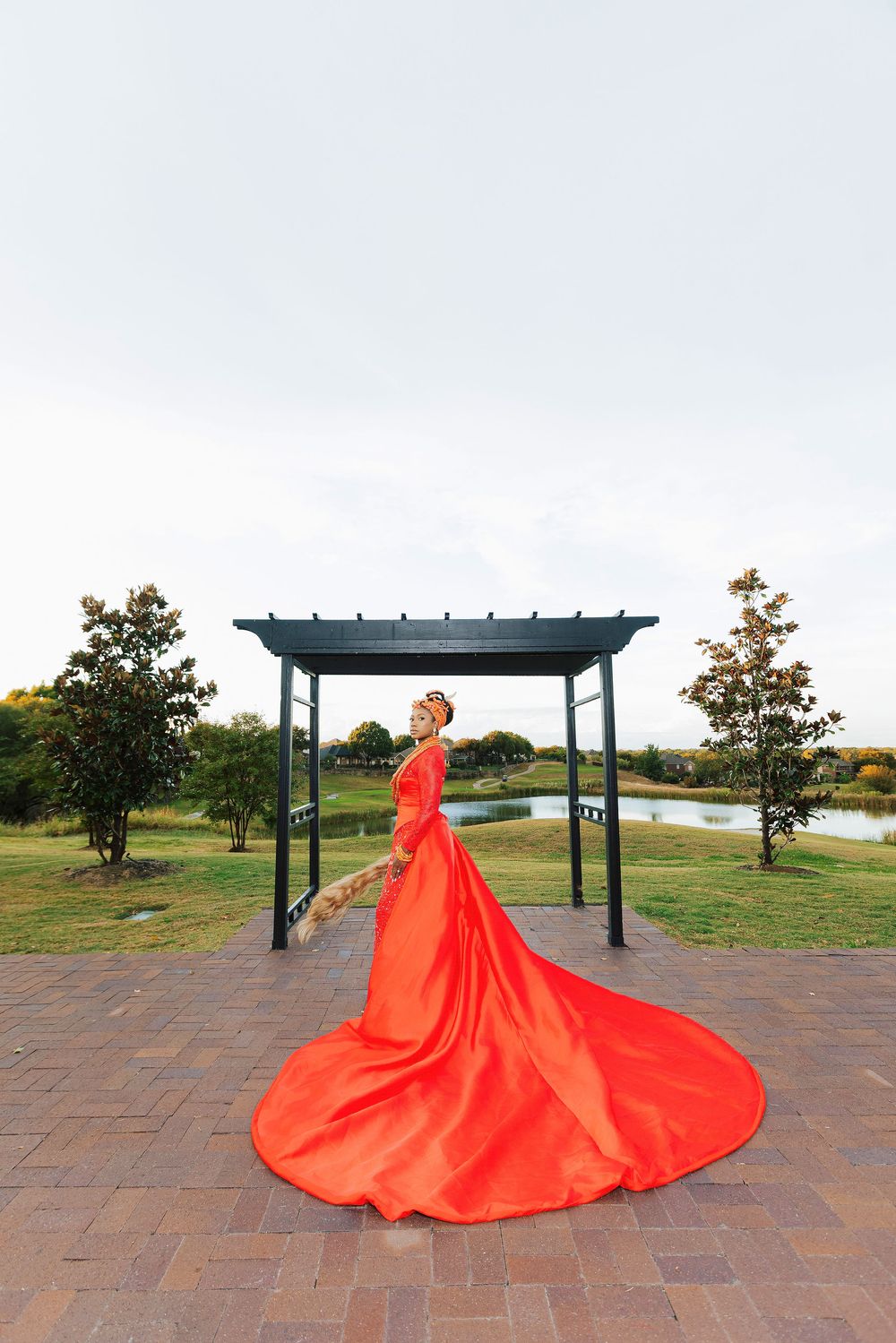 Woman in flowing red gown with long train poses under pergola in scenic park with pond backdrop.