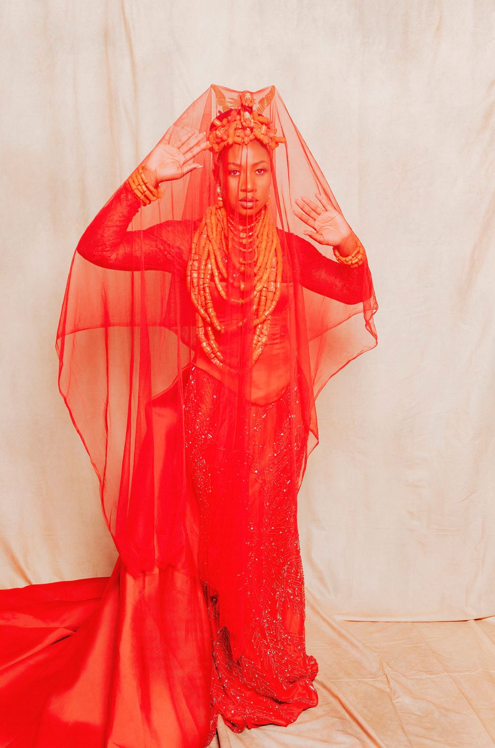 Woman in stunning all-red ensemble with beaded gown, veil, and headdress poses gracefully against neutral backdrop.