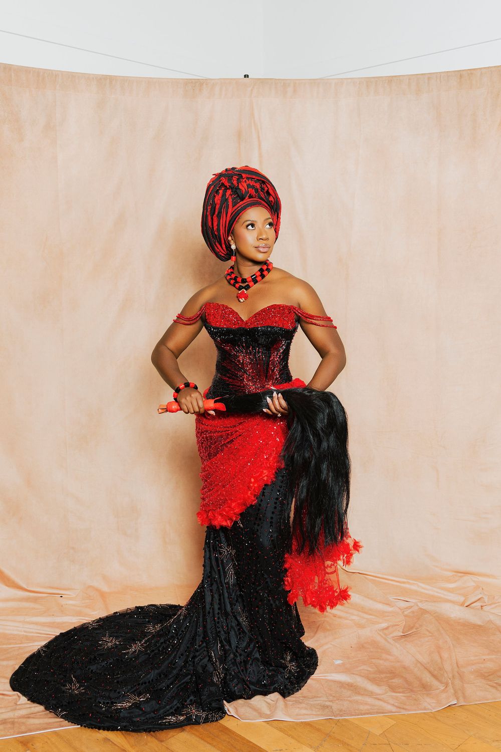 Woman in elegant red and black African traditional gown with gele headwrap and beaded jewelry posing gracefully.