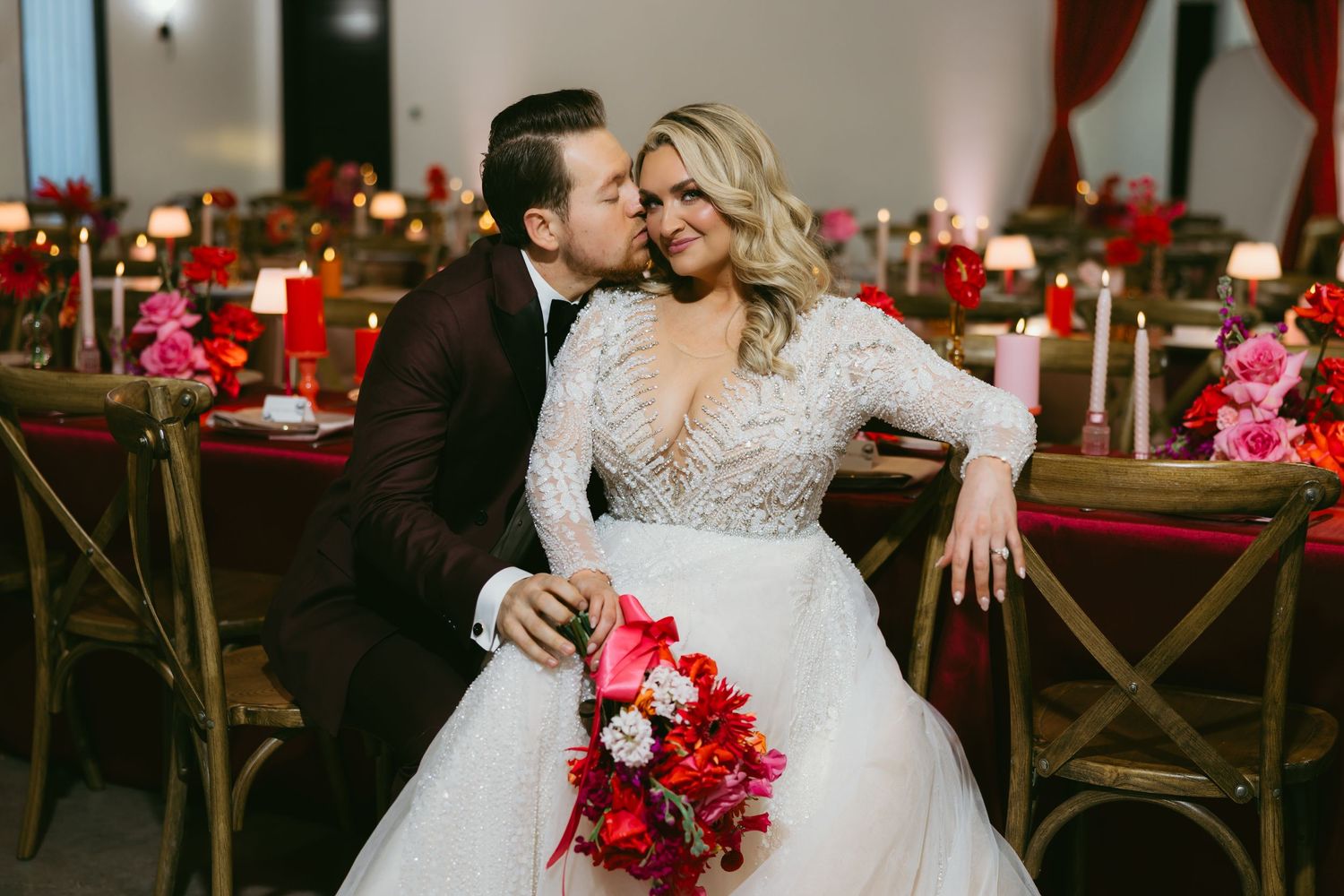 Bride and groom share a romantic kiss at a red-themed wedding reception with candles and floral centerpieces.
