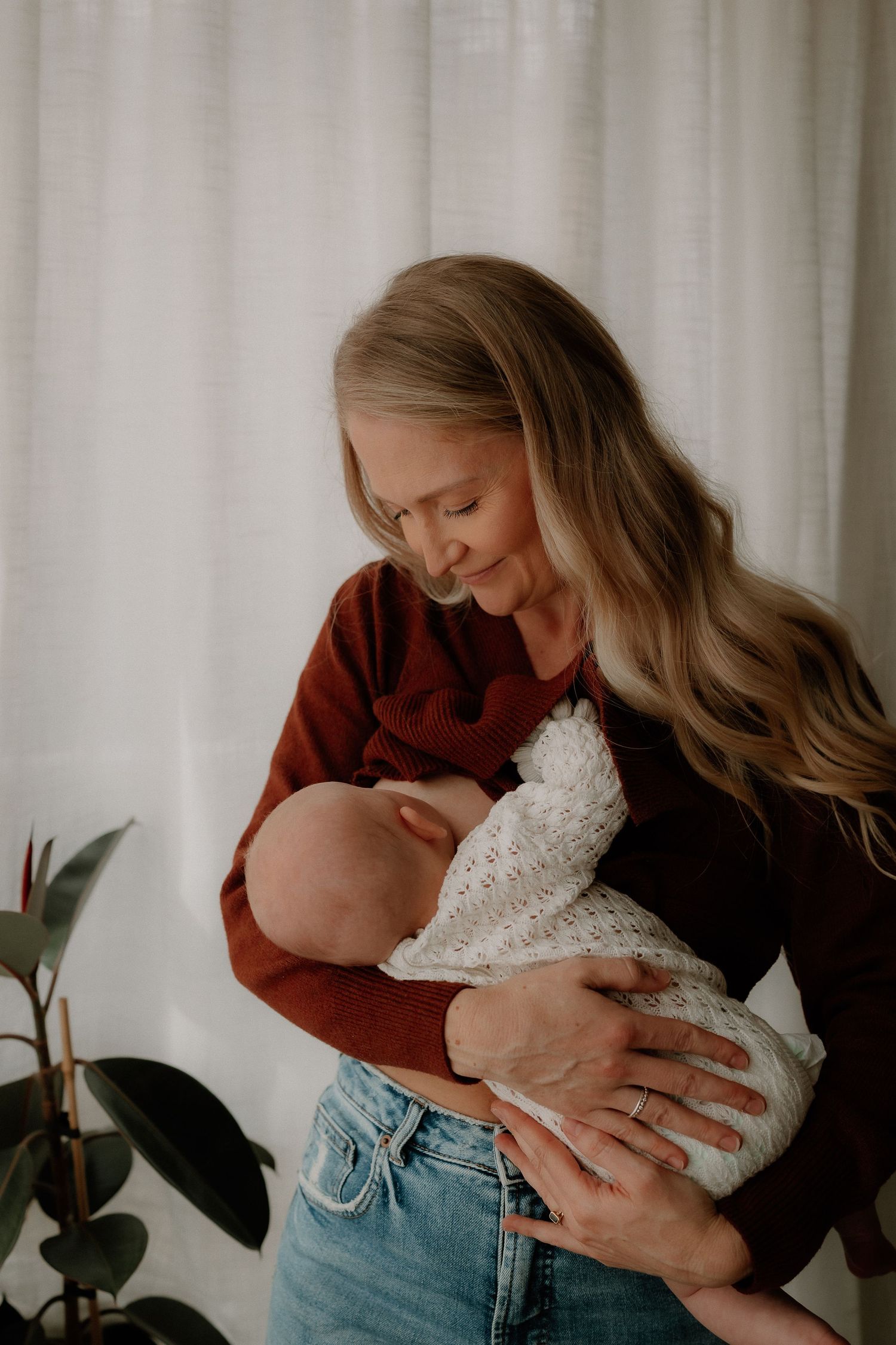 A woman with long blonde hair in a rust sweater and jeans tenderly holds a sleeping newborn wrapped in white knit.