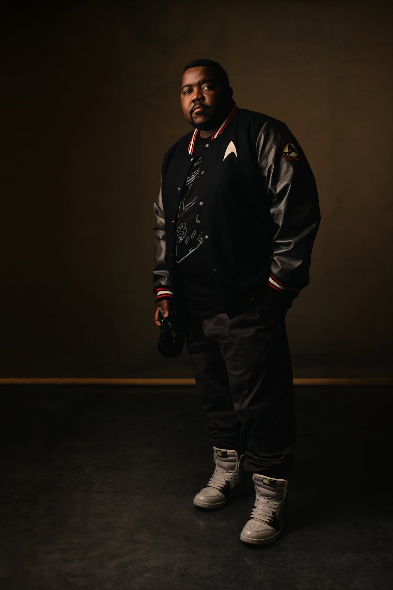 A heavyset man in a black varsity jacket and white sneakers poses confidently in a dark studio setting.