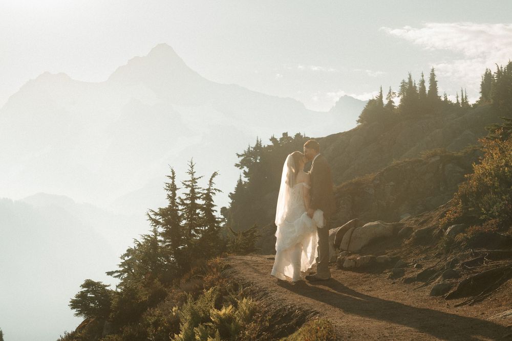 Couple in wedding attire embracing on a mountain trail with misty peaks and golden sunlight in the background.