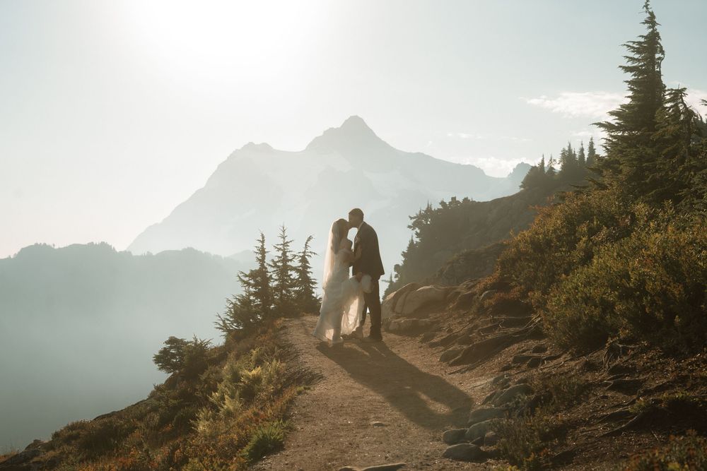 Couple shares a romantic kiss on a mountain trail with misty peaks and golden sunlight in the background.
