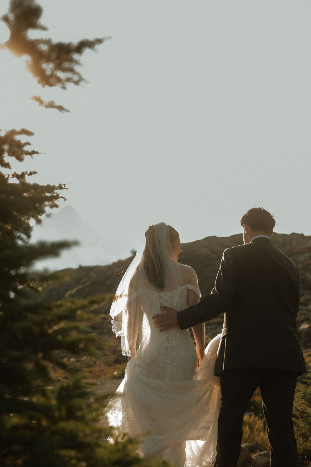 Bride and groom walking through mountain landscape at golden hour, bride in lace gown with veil.