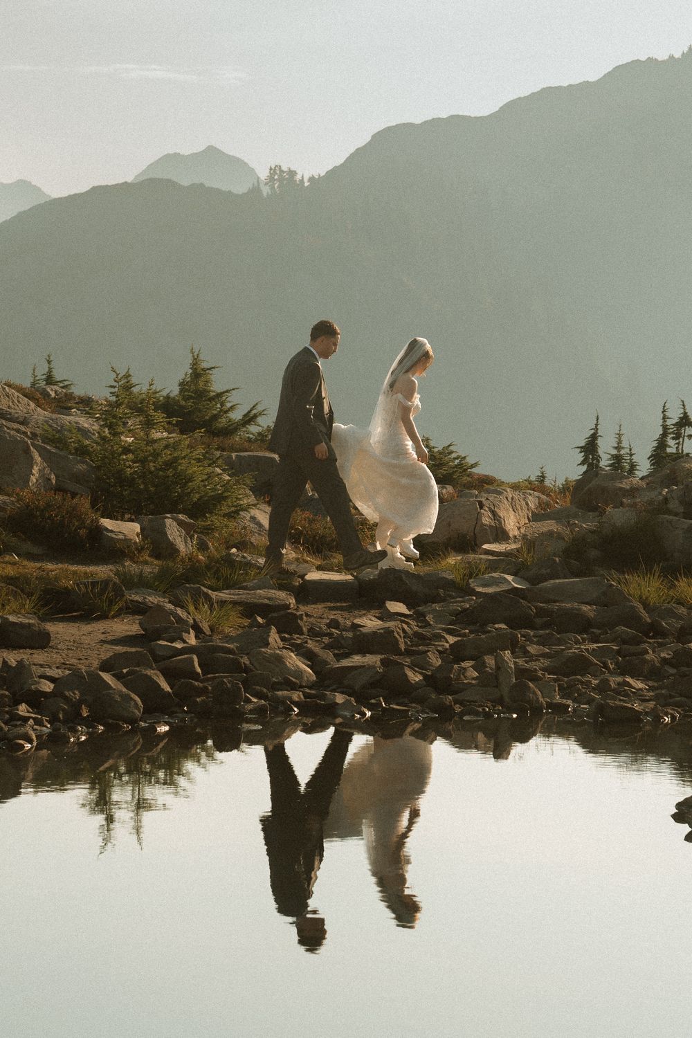 A bride and groom walk along a rocky alpine lake shore, their silhouettes reflected in calm water with misty mountains behind.