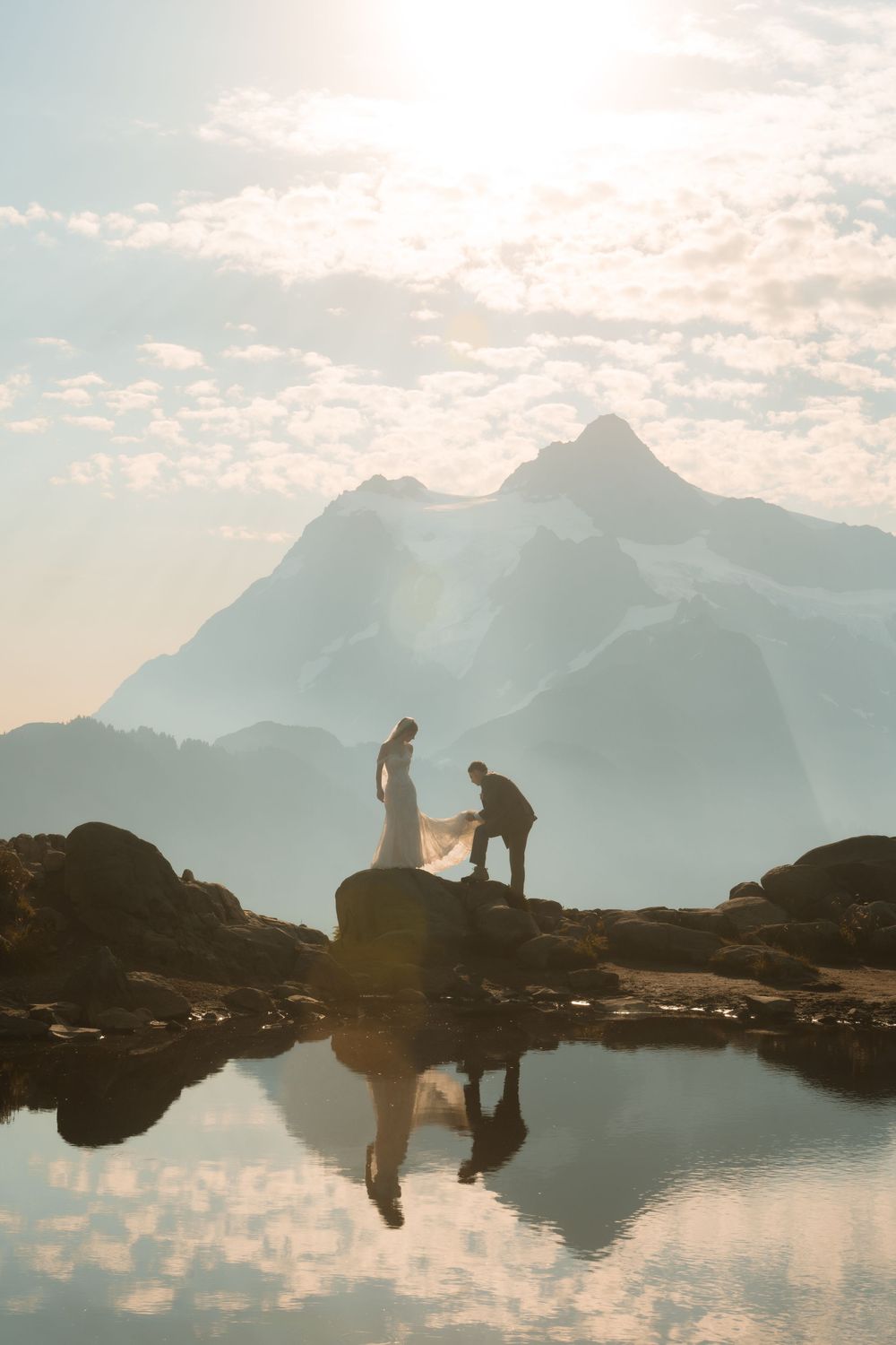 Couple in wedding attire standing on rocks by a reflective alpine lake with majestic mountain backdrop at golden hour.