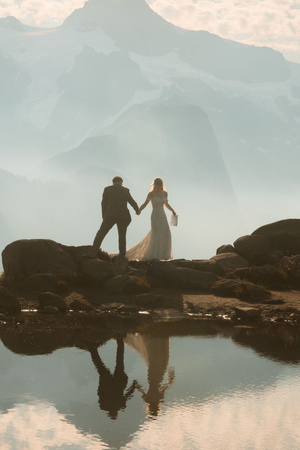 Bride and groom holding hands on rocky mountain shore, reflected in still water with misty peaks behind.