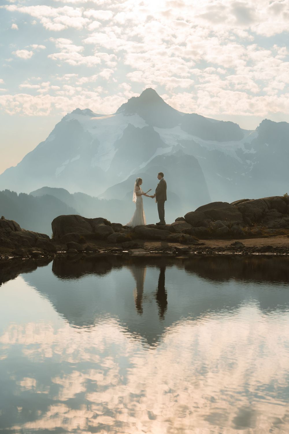 Wedding couple holding hands by alpine lake with mountain reflection, bride in white gown.