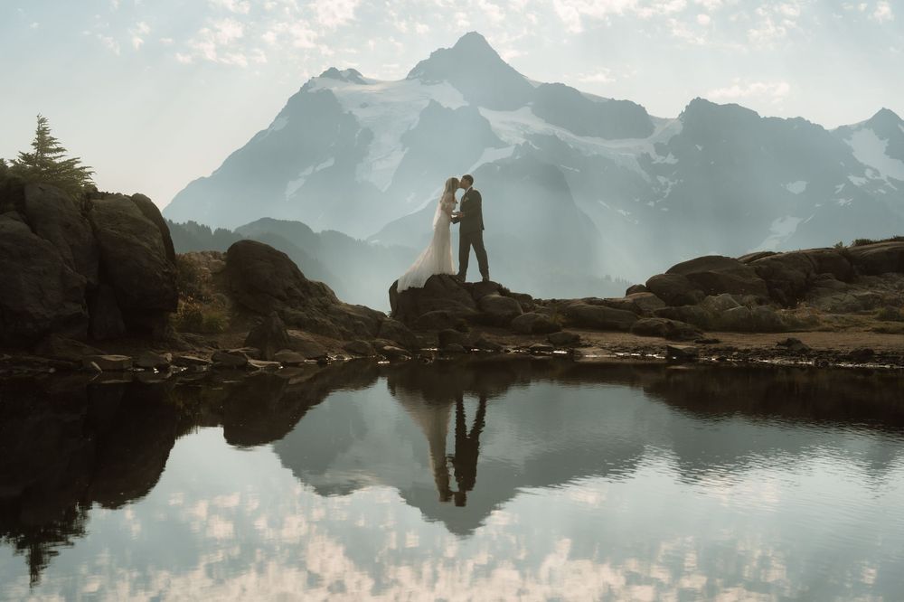 Bride and groom stand on rocky shore reflecting in alpine lake with misty mountain backdrop.