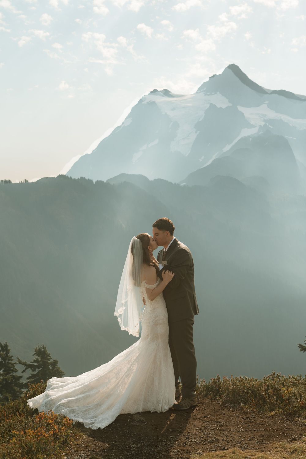 Bride and groom kiss on a mountain summit with a majestic snow-capped peak rising through misty clouds behind them.