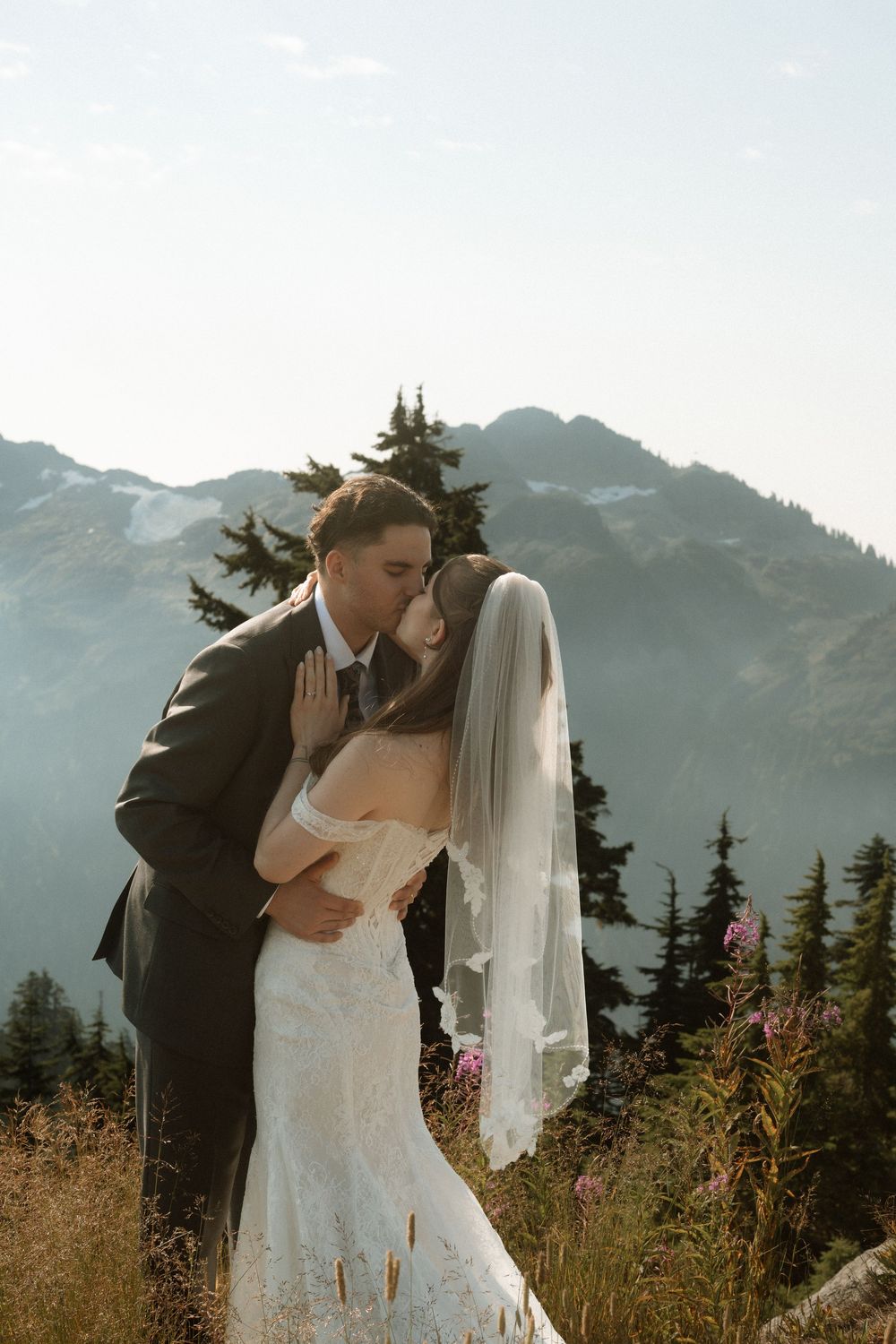 Bride and groom share a romantic kiss on a mountain overlook, with evergreen trees and snow-capped peaks in the background.
