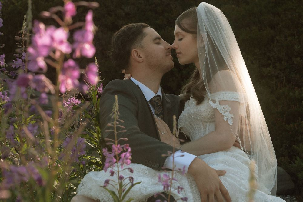 A bride and groom share a romantic kiss surrounded by purple flowers in a lush garden on their wedding day.
