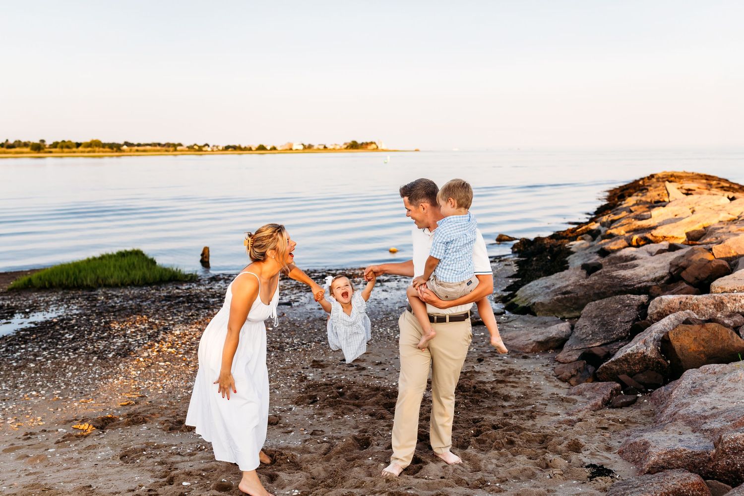 Family enjoying a golden hour beach moment, parents swinging toddler while holding young boy on rocky New England shore in Branford, CT