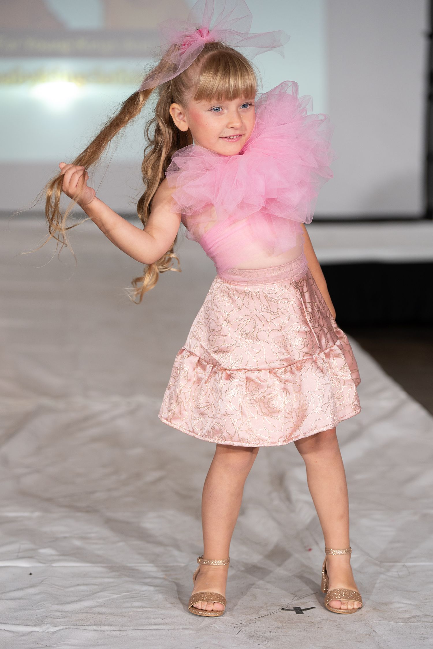Young girl models pink cape and black skirt on fashion runway, striking a confident pose in heels.