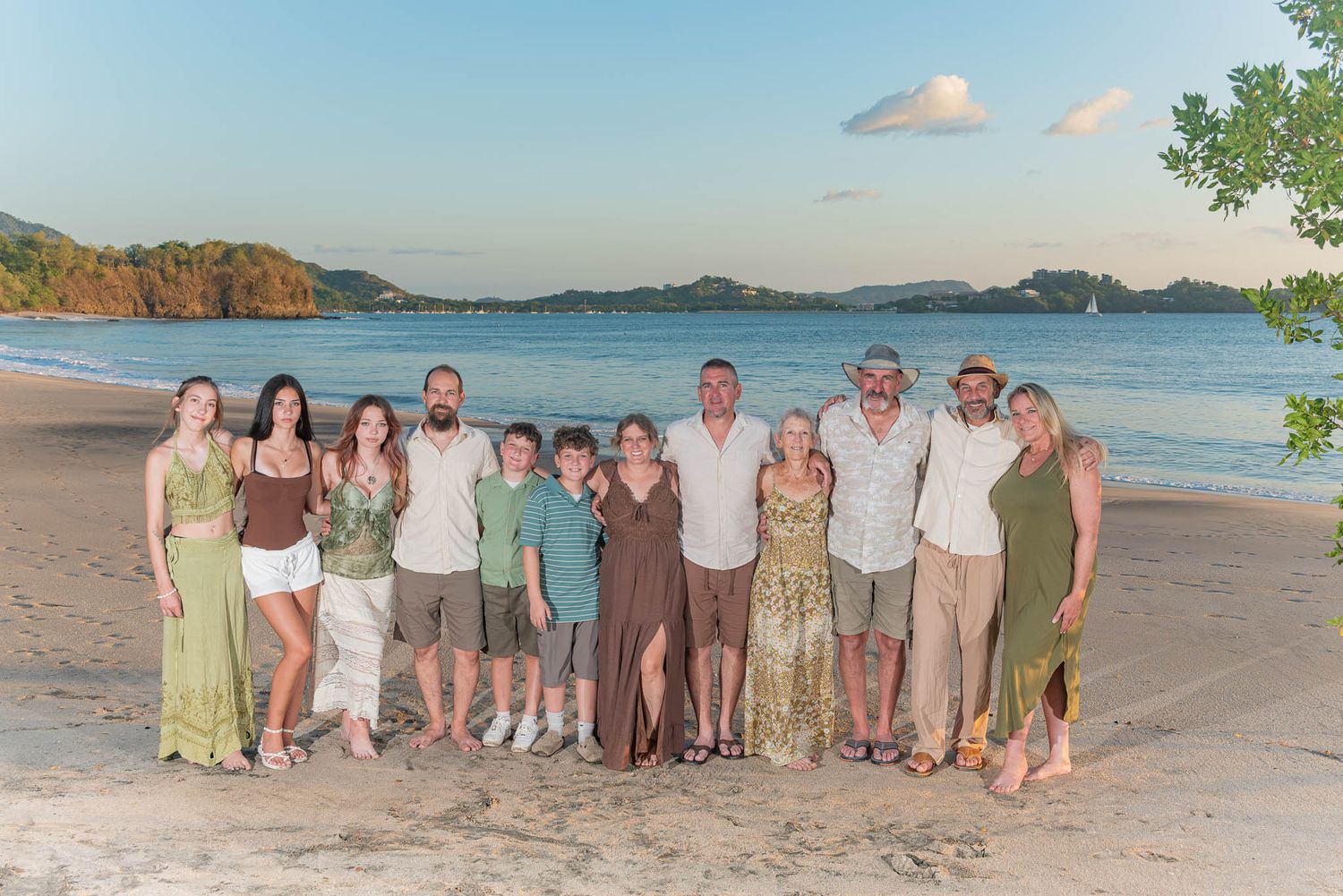 Large family group posing together on a tropical beach at sunset with calm blue water and lush hills in the background.