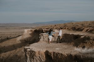 Couple standing on rocky desert overlook with vast arid landscape and mountains in the background.