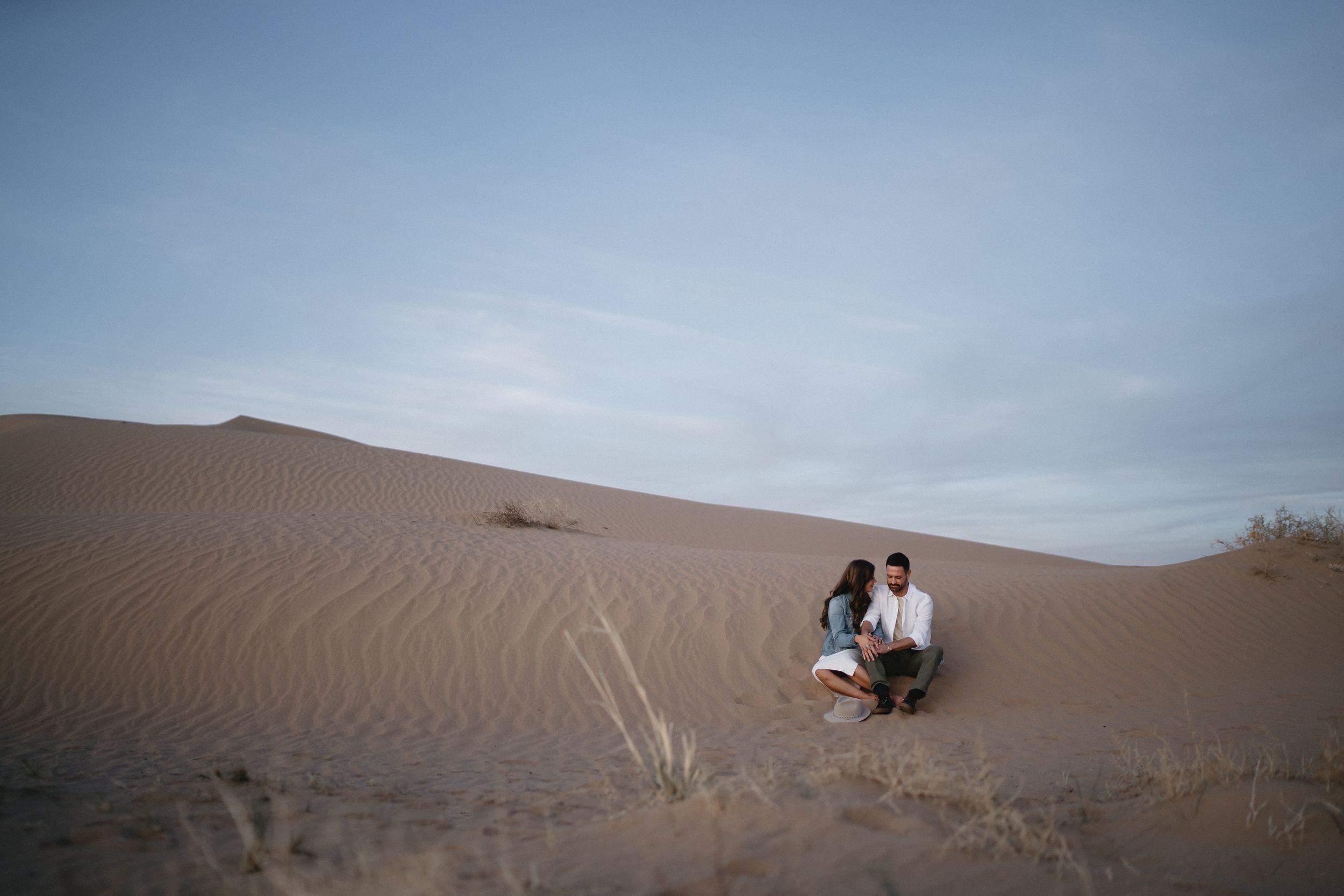 Couple sitting together on sandy desert dunes under a cloudy sky at dusk.