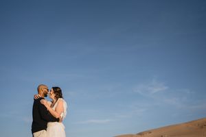Couple shares romantic moment in desert dunes under blue sky, bride in white gown and groom in black suit.
