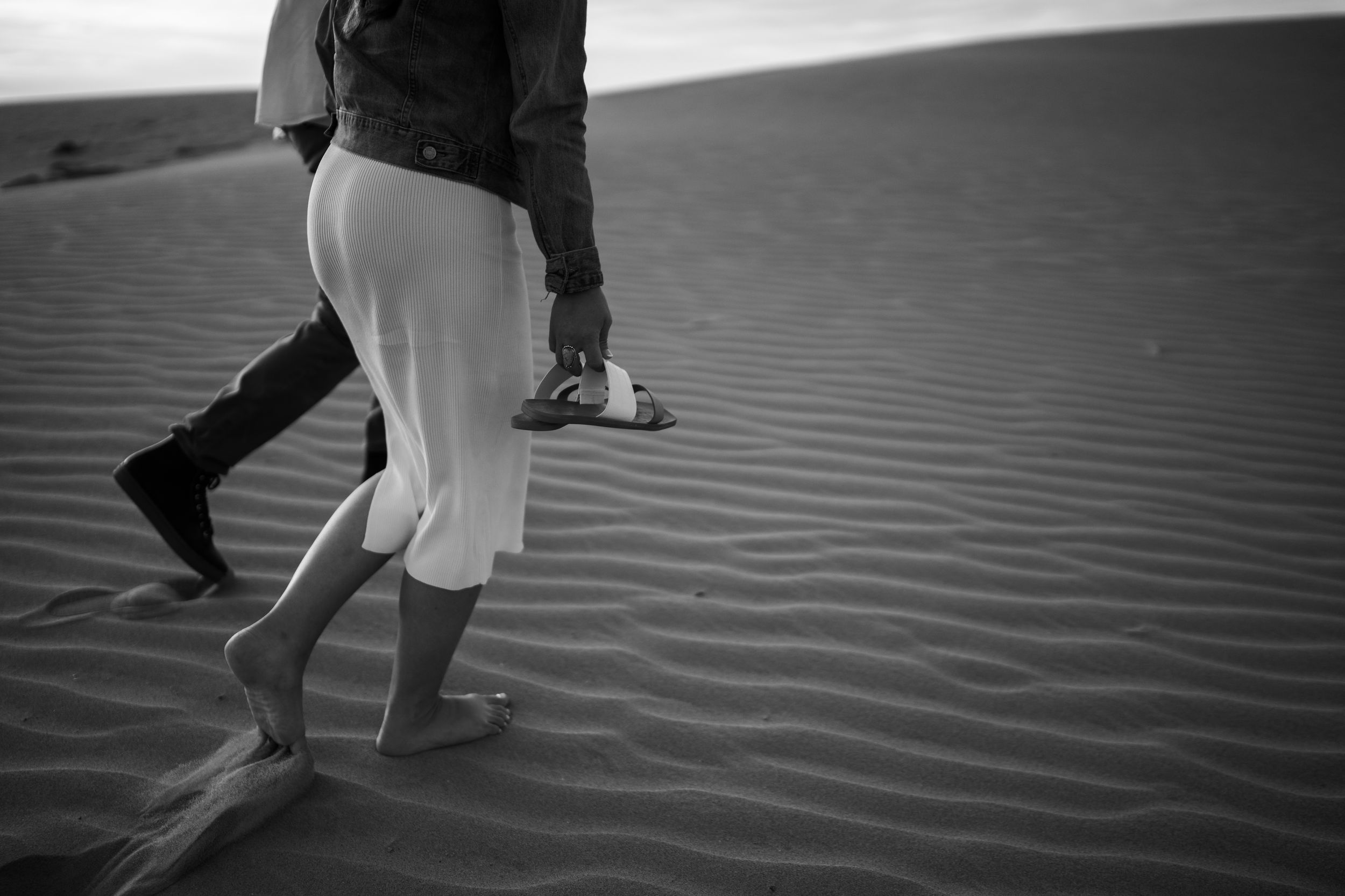 Black and white photo of a couple walking barefoot on sand dunes in a desert landscape.