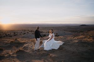 Couple in wedding attire holding hands at sunset in a desert landscape, bride's white gown flowing in the wind.