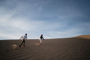 Couple running playfully on sandy desert dunes under a blue sky.