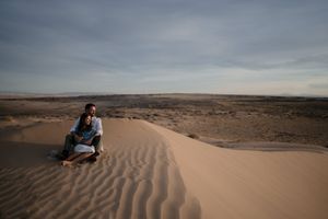 Couple sitting together on a sand dune in a vast desert landscape under a cloudy sky.