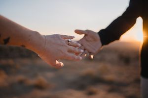 Two hands reaching toward each other at sunset, one wearing a ring, in a romantic desert engagement photo.