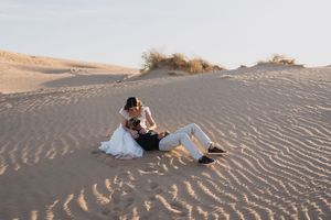 Bride and groom sitting together on sandy desert dunes during golden hour wedding photo shoot.