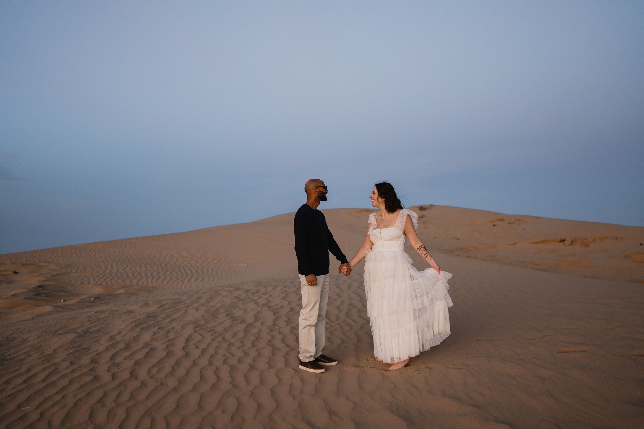 Couple in wedding attire holding hands on sandy desert dunes at dusk, bride in white gown, groom in black suit.