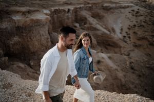 Couple holding hands walking on rocky desert canyon trail, man in white shirt, woman in denim jacket holding hat.